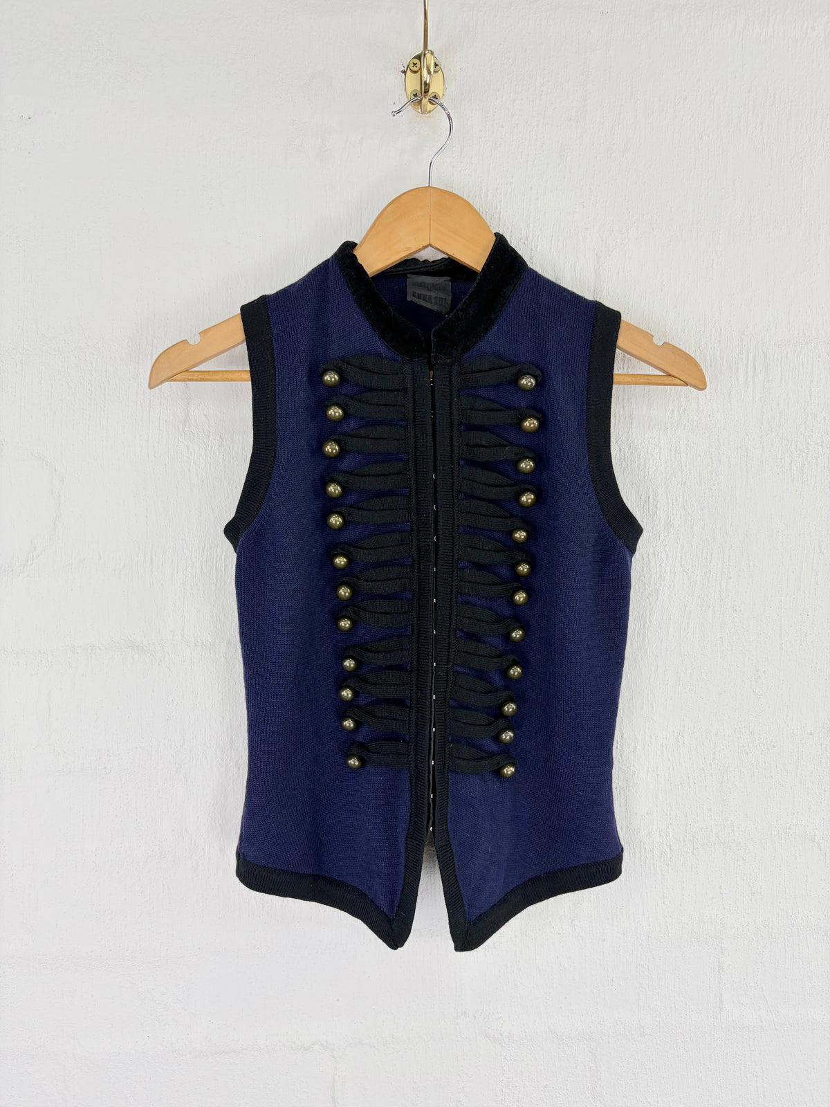 A navy blue sleeveless vest with black trim and decorative gold-tone military-style buttons, hanging on a wooden hanger against a white textured wall.