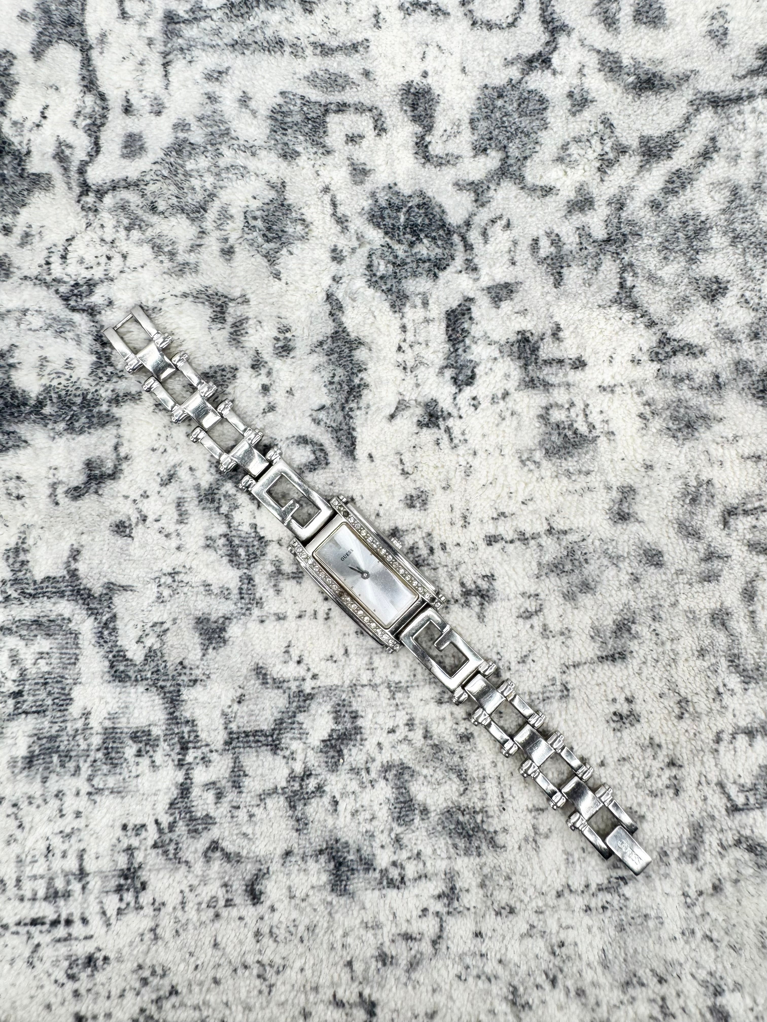 A silver-toned metal link bracelet watch with a rectangular center section, photographed diagonally on a textured gray and white patterned rug.