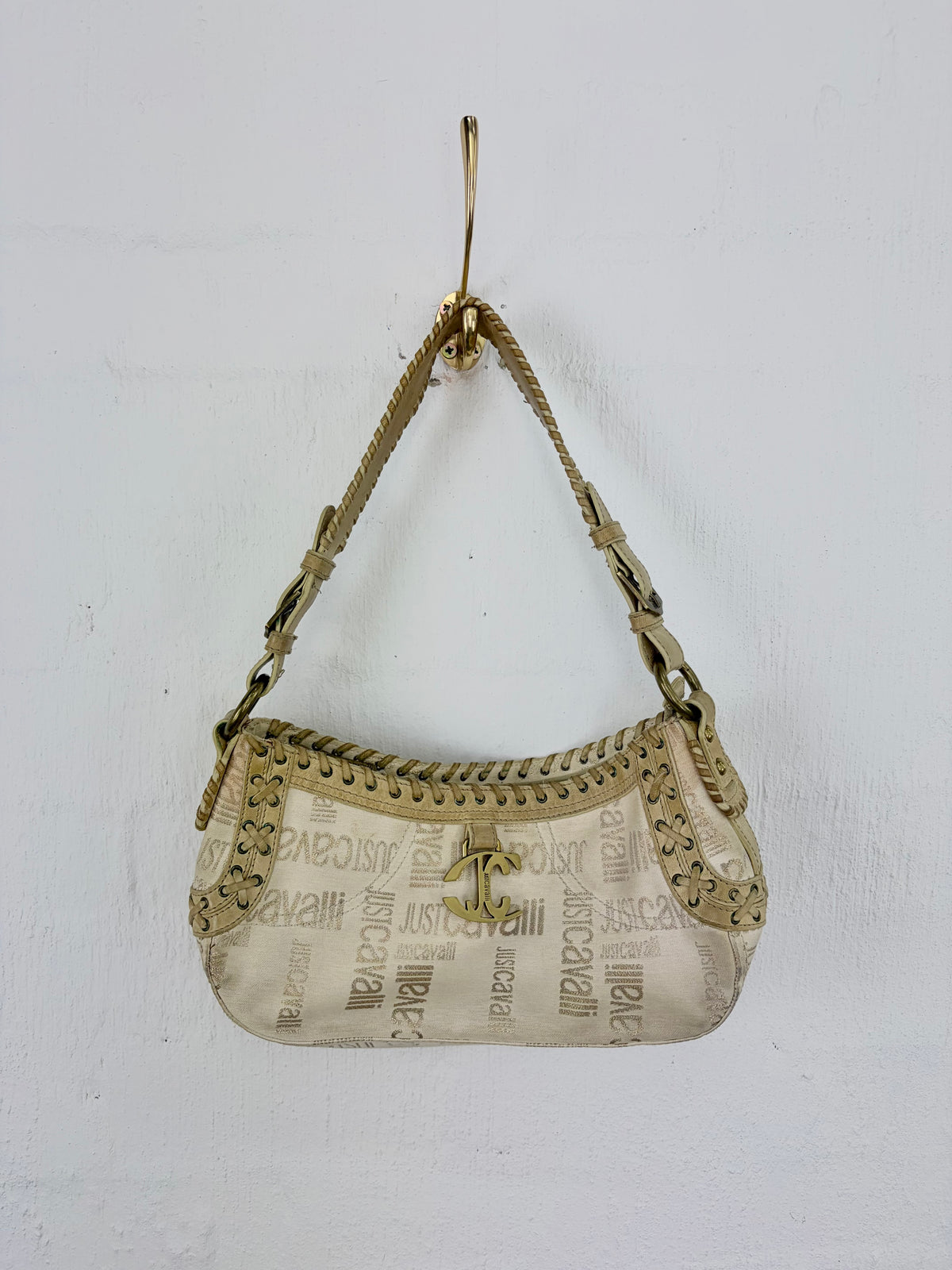 A vintage Chanel hobo bag in beige and gold-tone monogram canvas, hanging from a gold hook against a white wall. The bag features a braided shoulder strap, gold-tone studs, and the iconic interlocking CC logo.