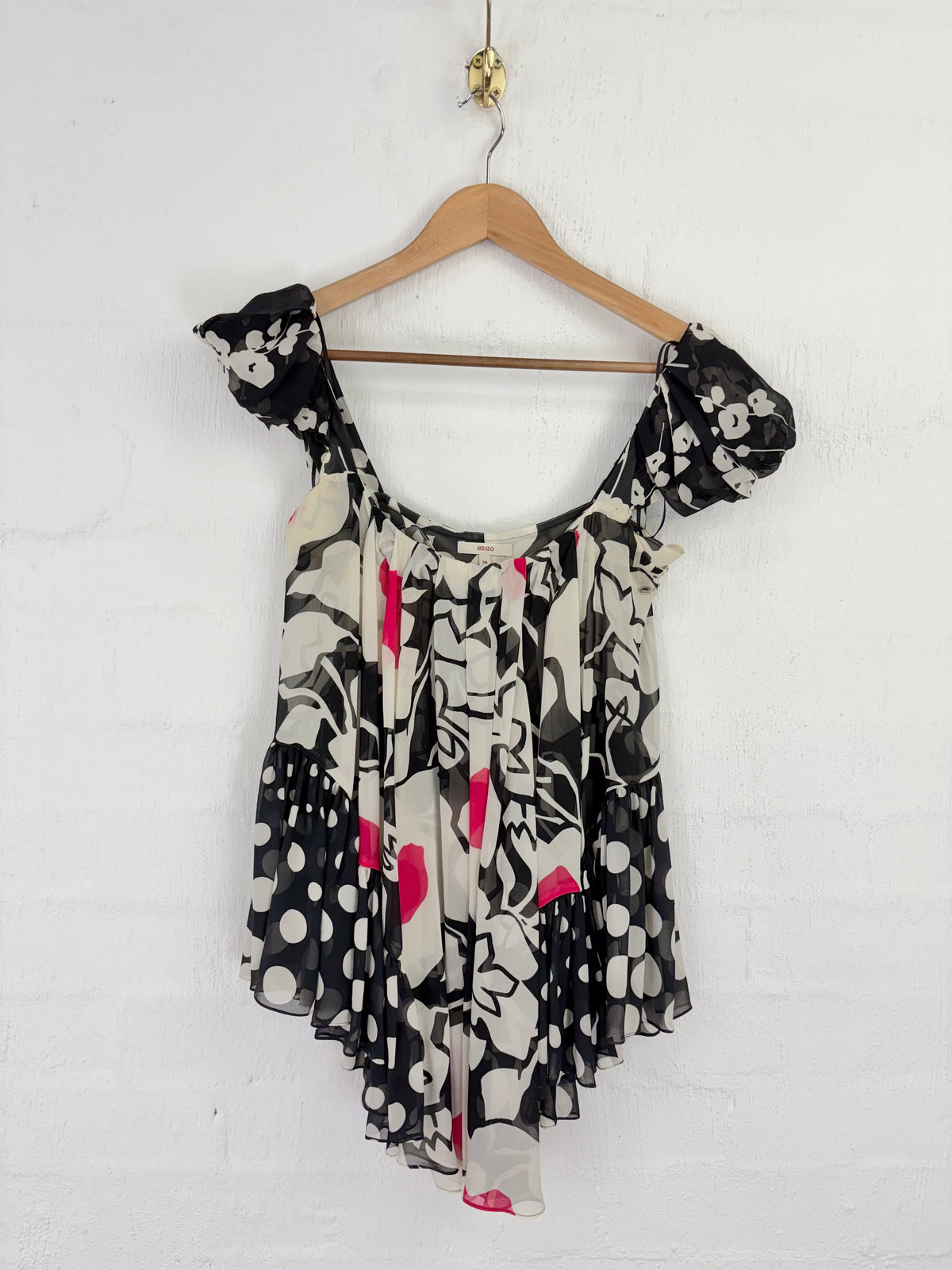 A black, white, and pink abstract print top with ruffled cap sleeves, hanging on a wooden hanger against a white painted brick wall.