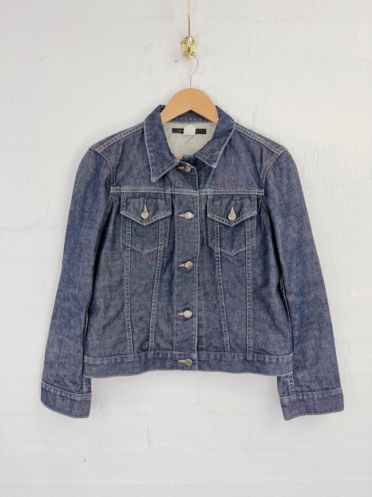 A blue denim jacket hanging on a wooden hanger against a white painted brick wall.