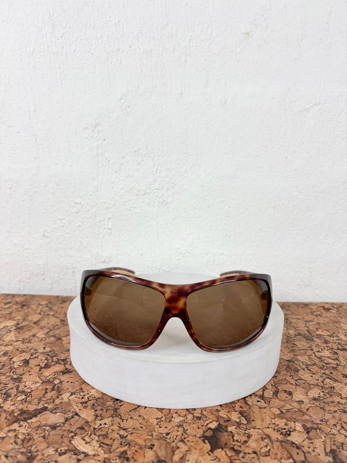 Tortoiseshell sunglasses with brown lenses displayed on a white circular stand over a cork surface against a white wall.