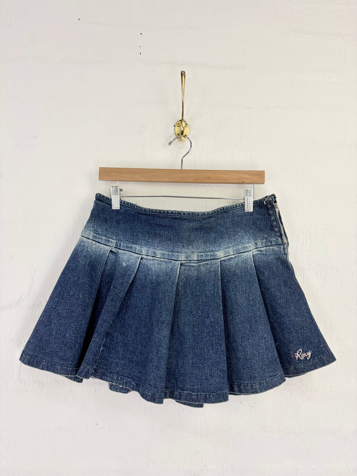A blue denim pleated mini skirt with a faded wash and a small white embroidered logo on the lower right hem, hanging from a wooden hanger against a white wall.