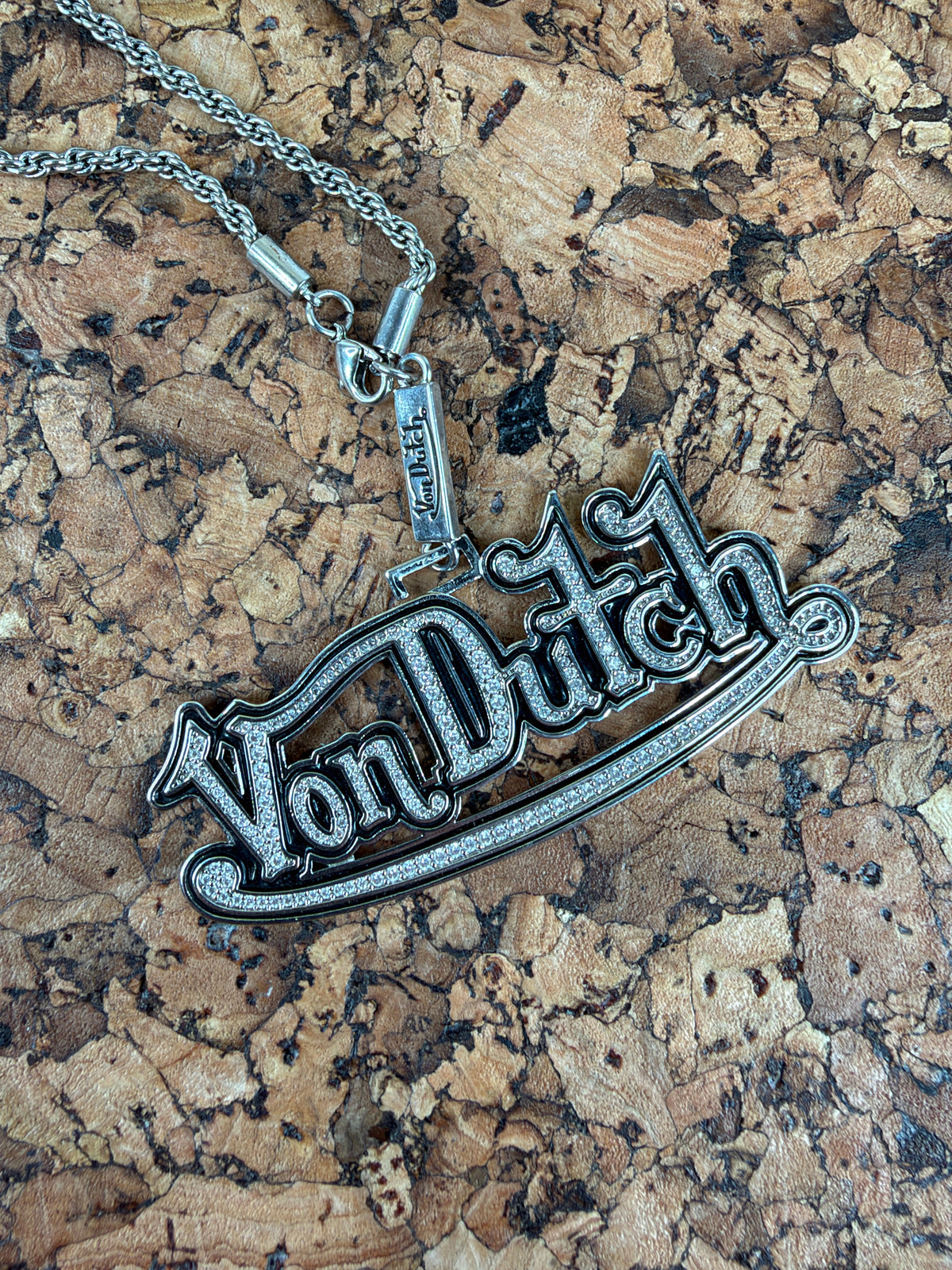 A silver-toned gothic-style pendant necklace with the word 'Vanity' in stylized lettering, displayed on a textured cork background.