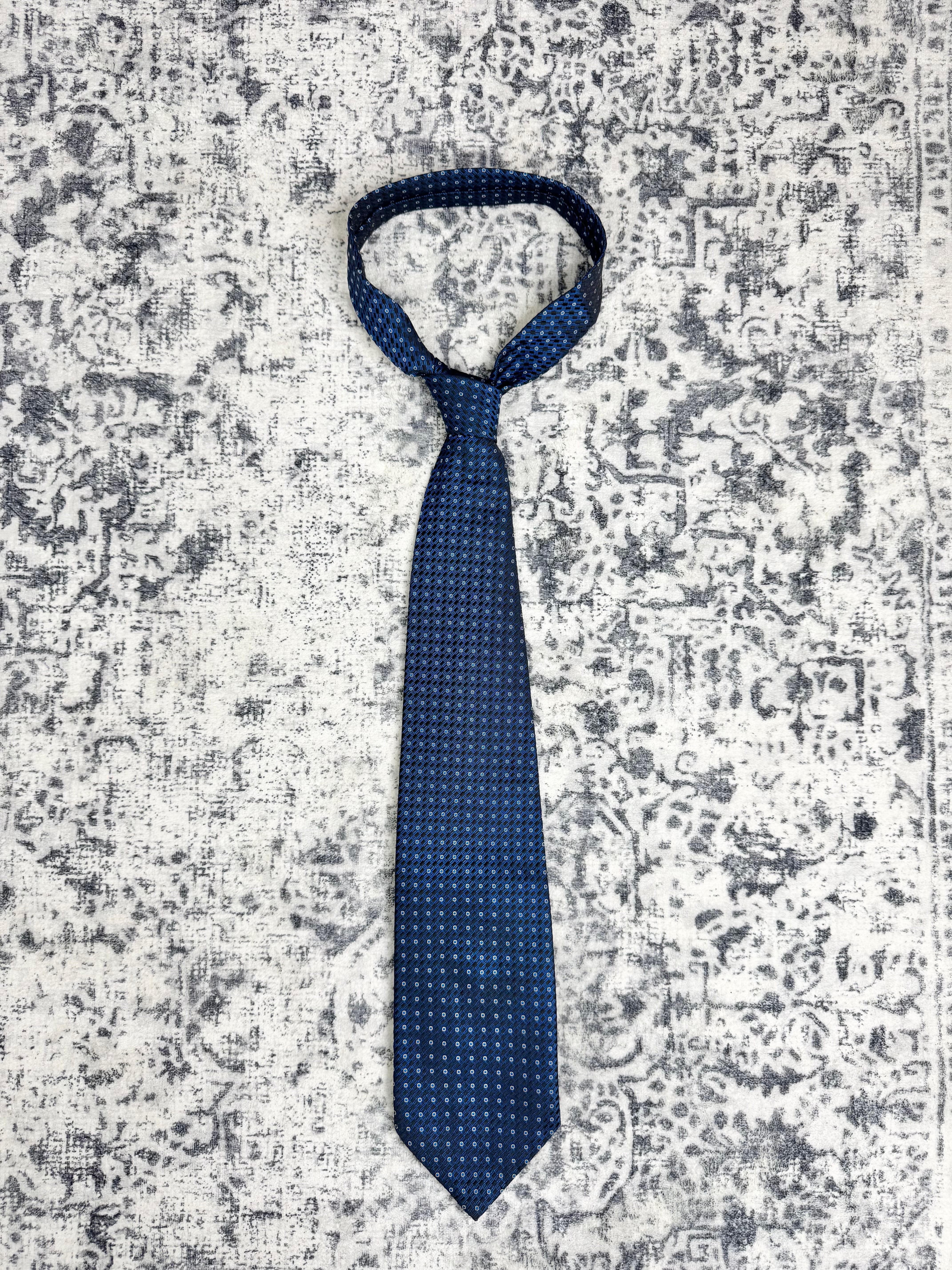 A dark blue necktie with a small white geometric pattern, tied in a standard knot and laid flat on a distressed gray and white patterned rug.