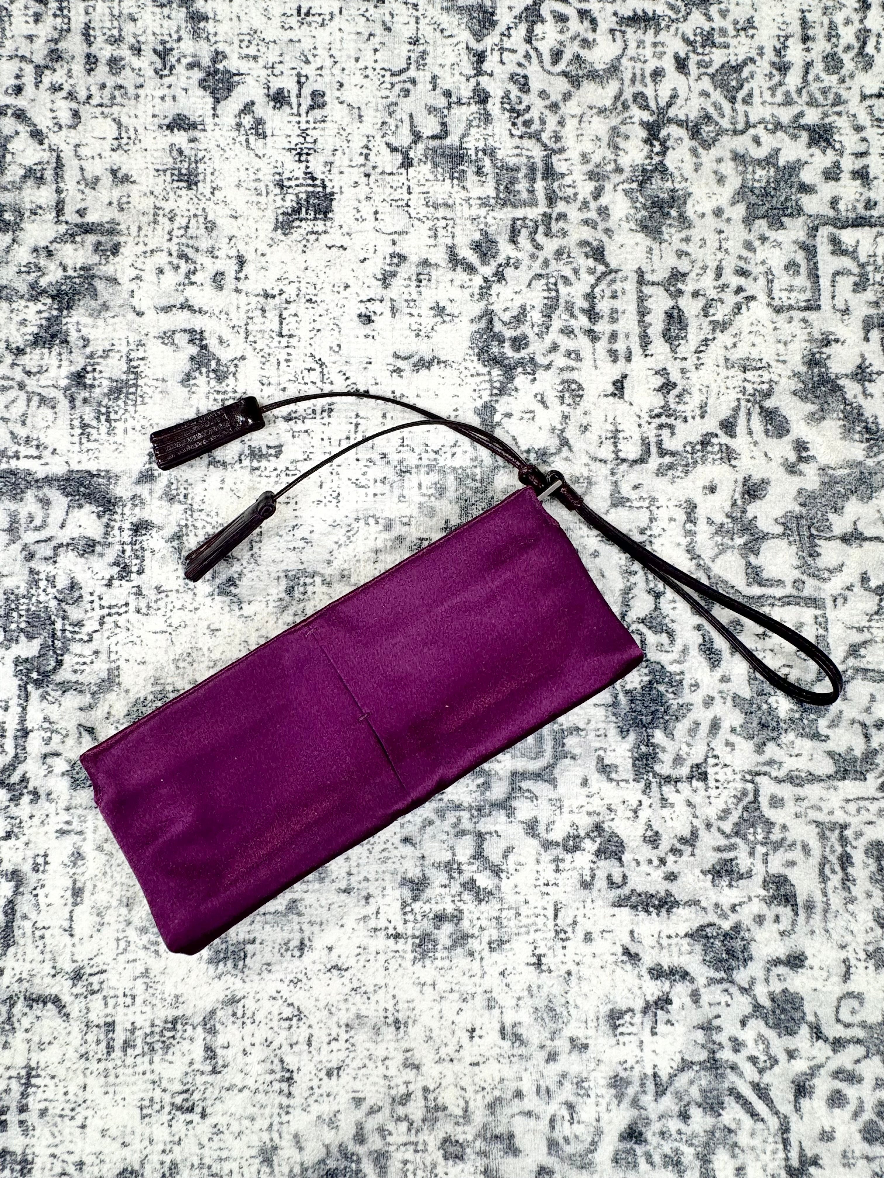 A purple rectangular fabric pouch or sleeve with a black wrist strap and black tassel-like ties, laid flat on a distressed gray and white patterned rug.