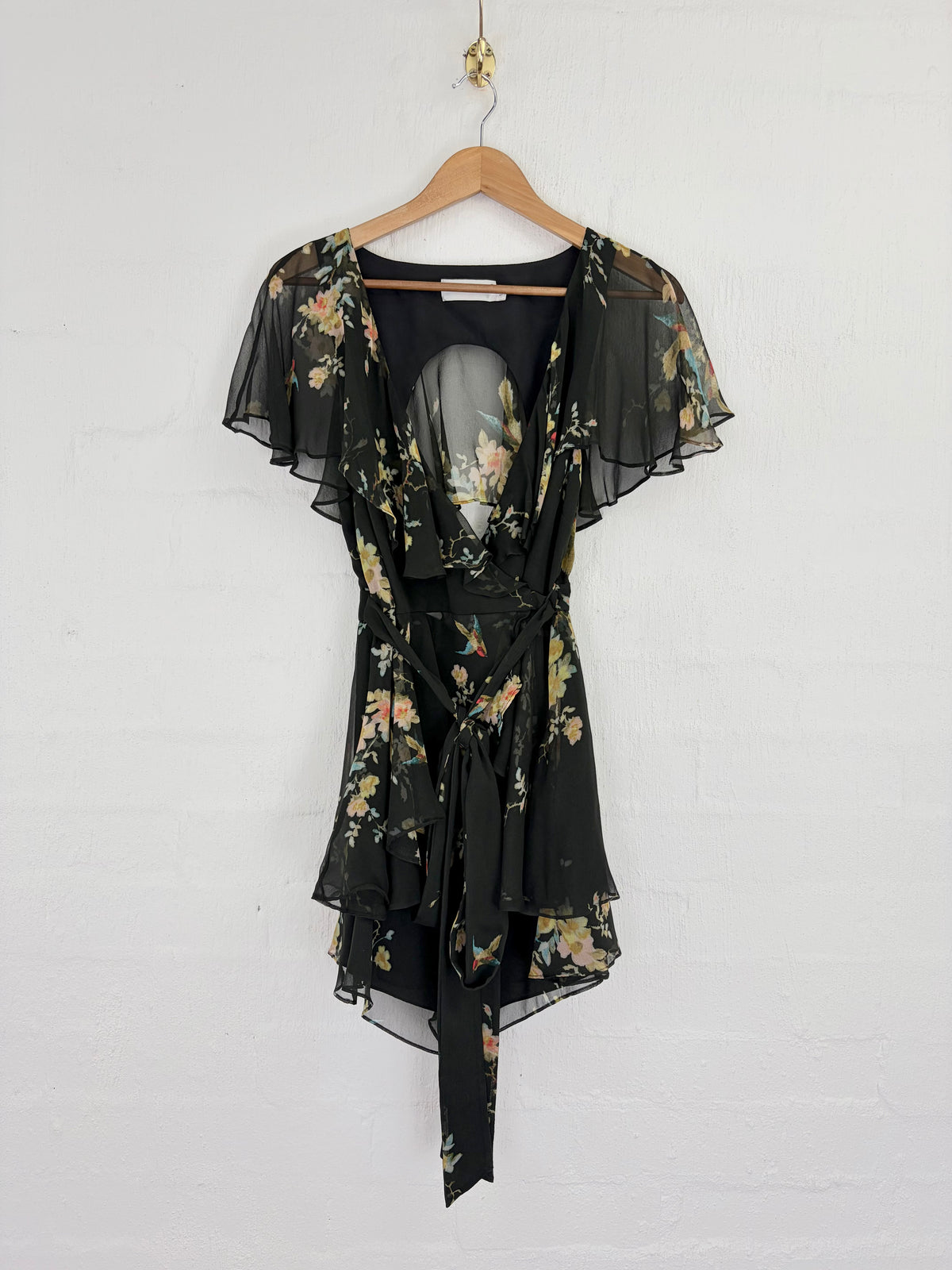 A black sheer wrap dress with a floral print, hanging on a wooden hanger against a white brick wall.
