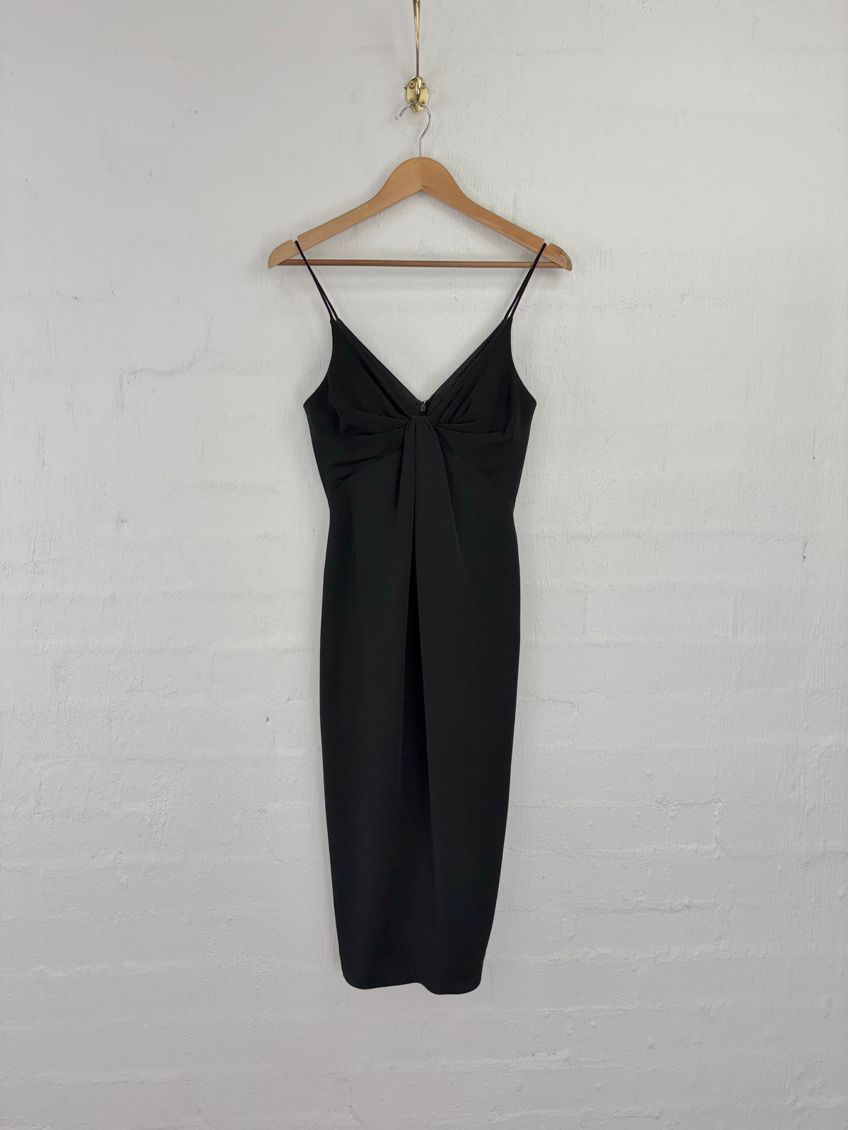 A black spaghetti-strap midi dress with a twisted knot detail at the bust, hanging from a wooden hanger against a white painted brick wall.