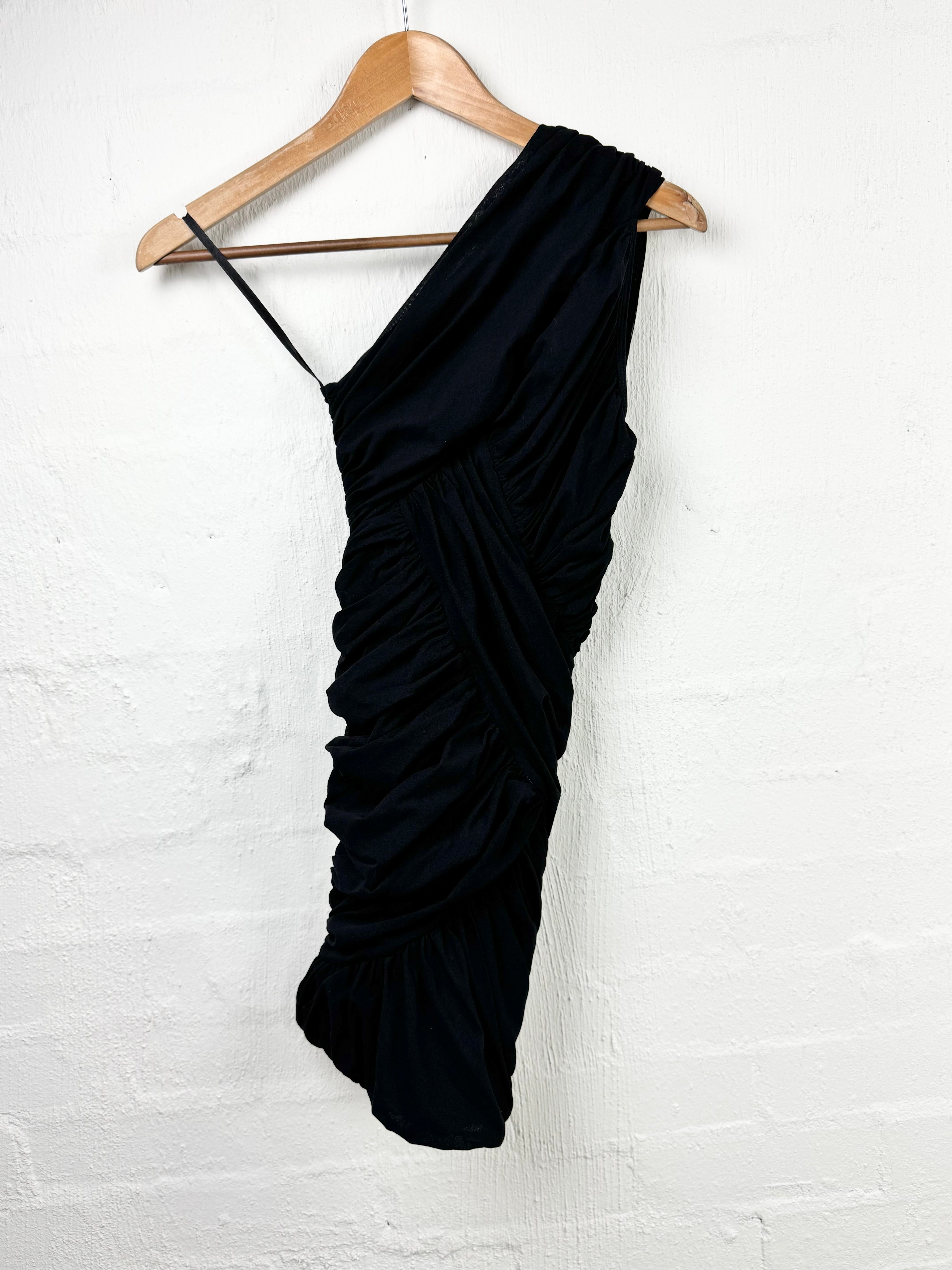 A black one-shoulder ruched dress hanging from a wooden hanger against a white painted brick wall.
