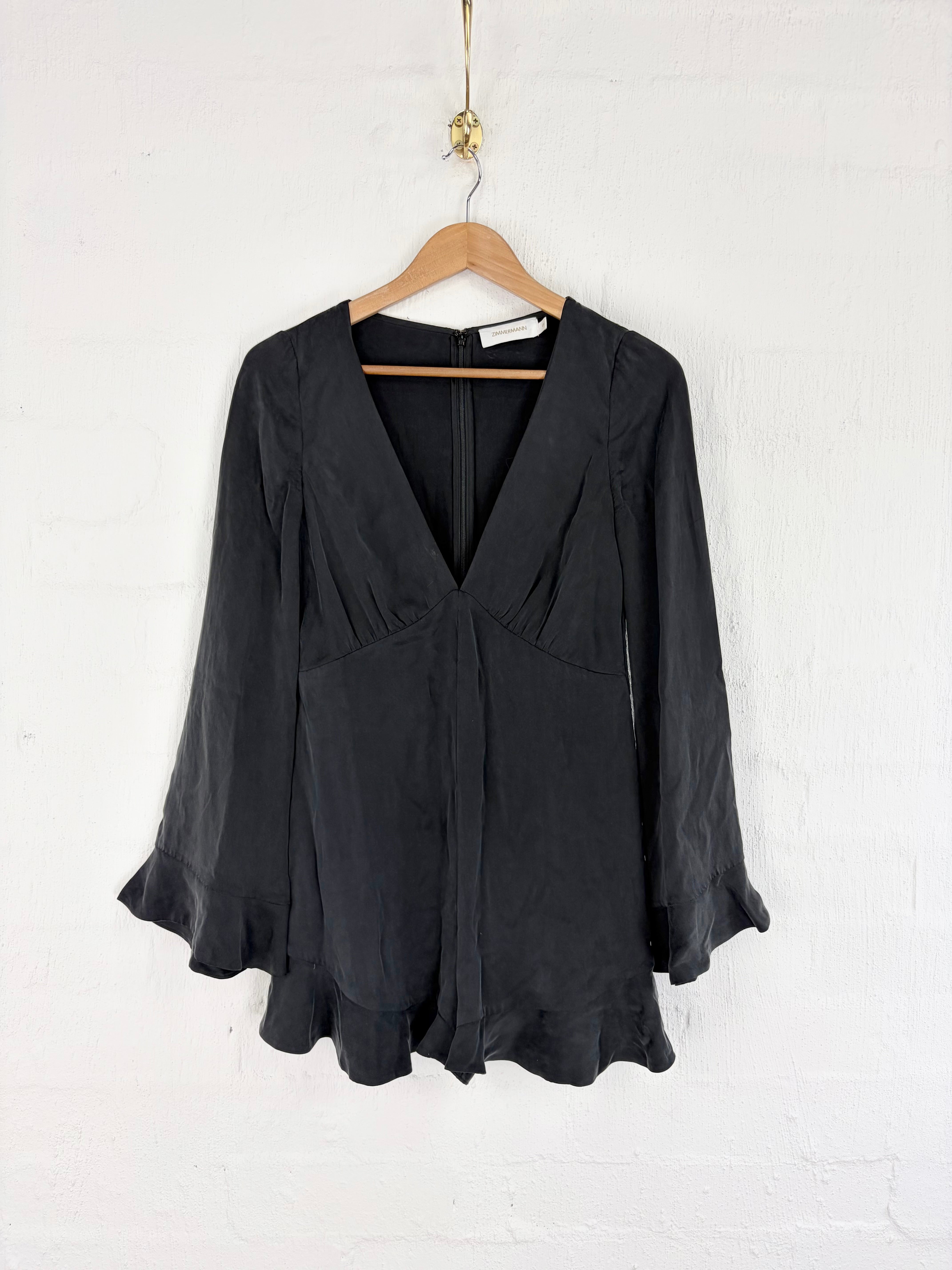 A black long-sleeve blouse with a deep V-neckline and gathered front detail, hanging on a wooden hanger against a white painted brick wall.