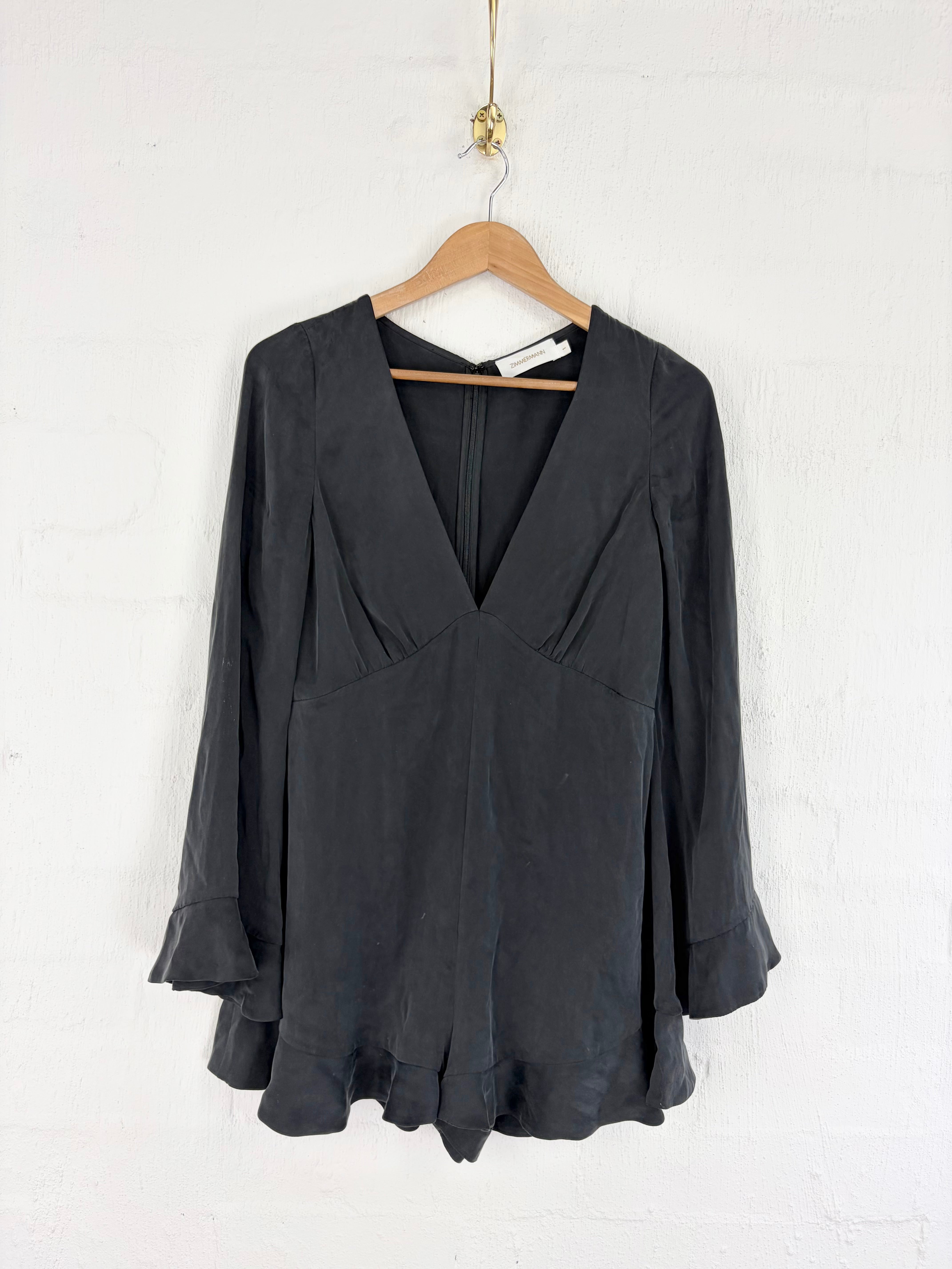 A black long-sleeve blouse with a deep V-neckline and ruffled cuffs, hanging on a wooden hanger against a white painted brick wall.