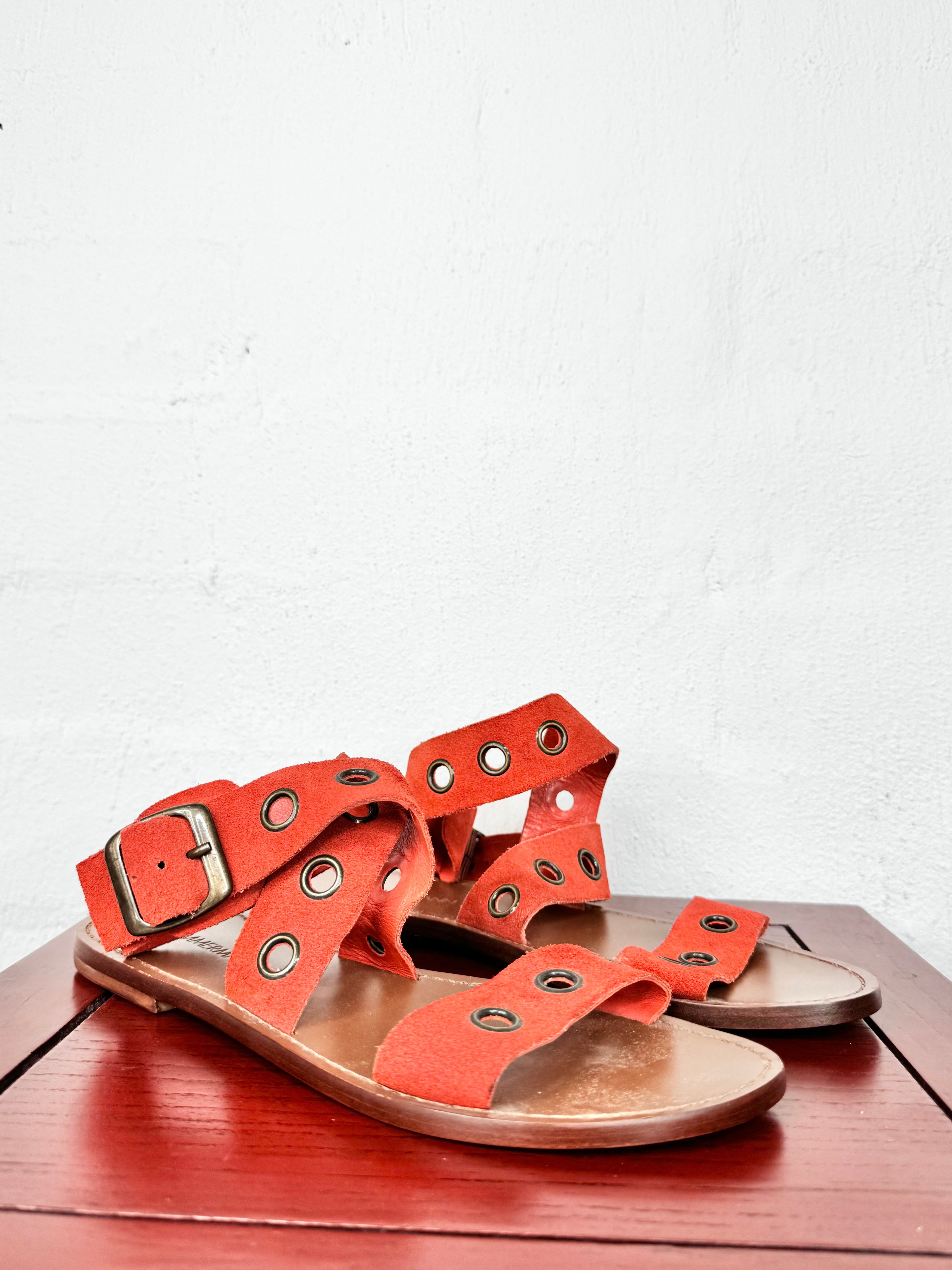 A pair of orange-red leather flat sandals with multiple cutout straps, metal eyelets, and a buckle, displayed on a reddish-brown wooden surface against a white wall.