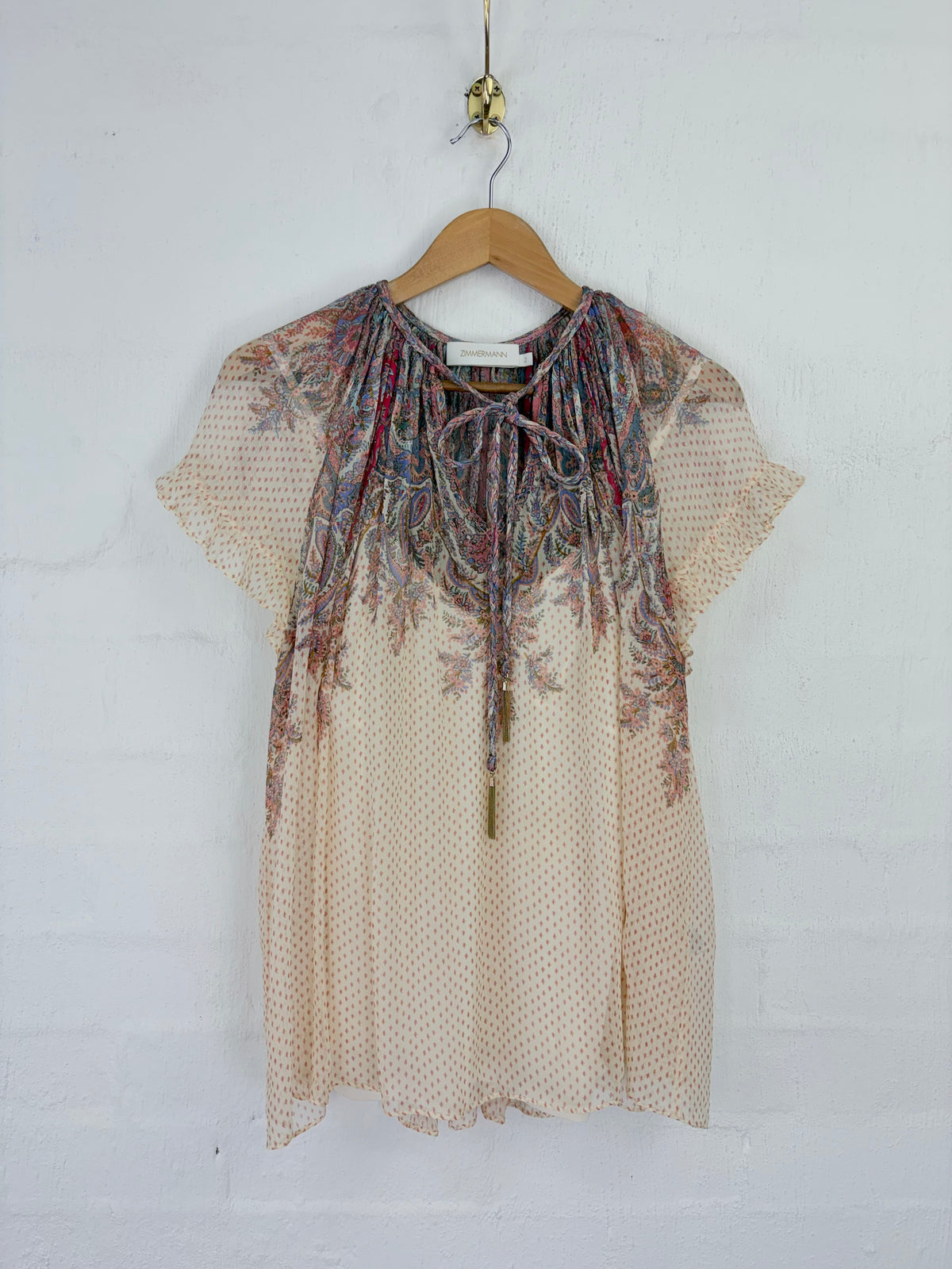 A short-sleeve blouse with a sheer beige mesh body and a heavily embroidered floral yoke in muted red, blue, and brown tones, hanging on a wooden hanger against a white painted brick wall.