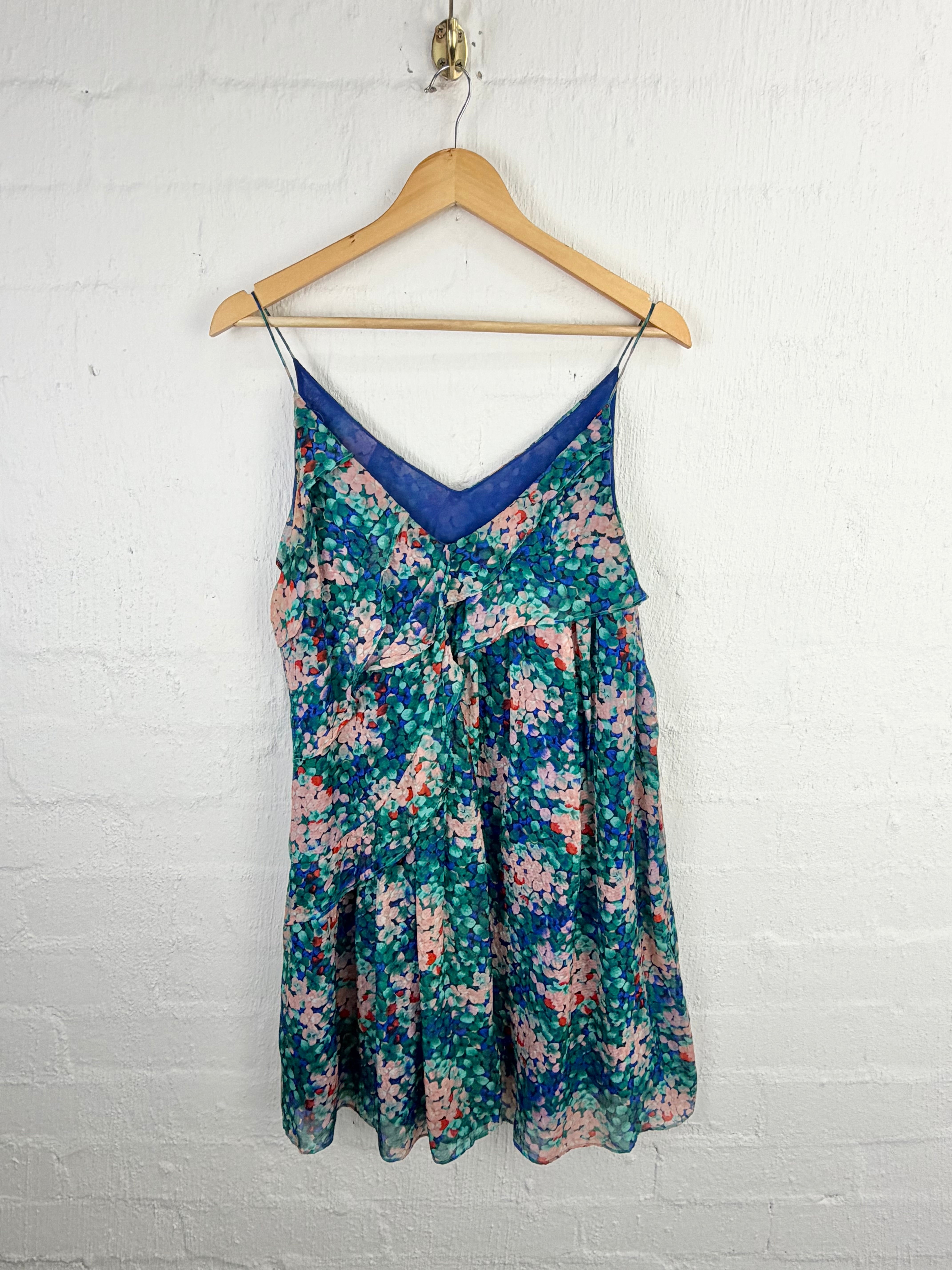 A sleeveless V-neck slip dress with a blue mesh trim and a green, pink, and blue floral print, hanging on a wooden hanger against a white painted brick wall.