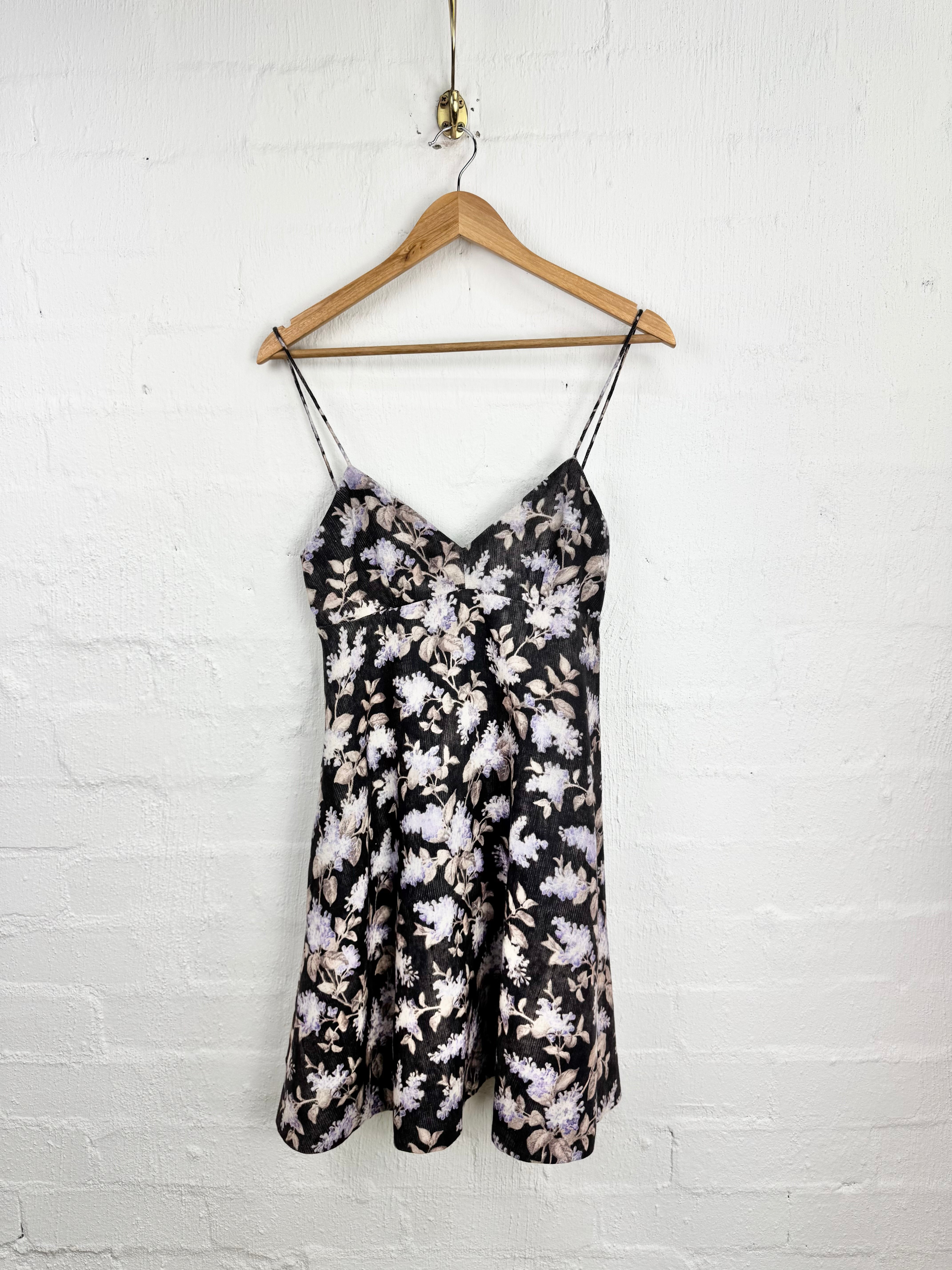 A black floral slip dress with thin spaghetti straps and a gathered sweetheart neckline, hanging on a wooden hanger against a white painted brick wall.