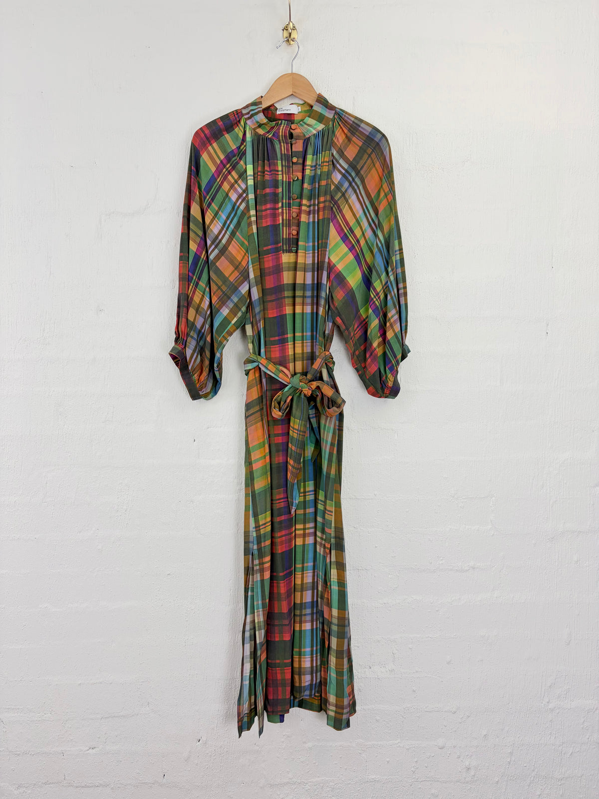 A long-sleeve maxi dress with a multicolor plaid pattern, featuring a tie waist belt and a button-up front, hanging on a wooden hanger against a white brick wall.